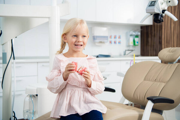 A portrait of a little female patient sitting in the dentists chair with dentures in her hand and smiling with a toothy smile