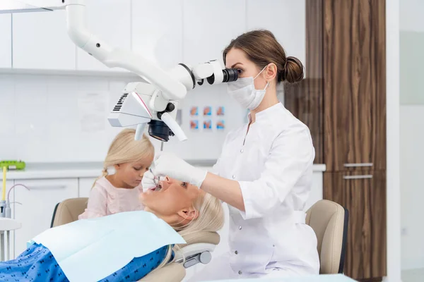 Um dentista olhando para uma boca de mulher através de um microscópio dental, a filha de mulher está ao lado de sua mãe, ela está entediada, esperando por ela para completar o check-up — Fotografia de Stock