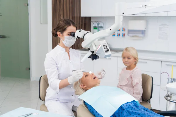 A blond woman having her teeth checked at the dentists office, with her daughter waiting for her — Stock Photo, Image