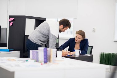 Bir iş kadını masasında otururken çalışanına bir şey anlatıyor. Çalışan dikkatlice dinliyor, öne eğiliyor.