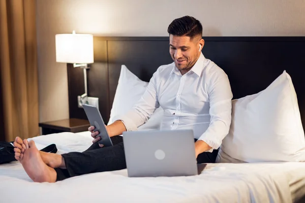 Formalite icabı giyinmiş genç ve mutlu bir iş adamı otel odasında yatağında oturmuş gülümsüyor.