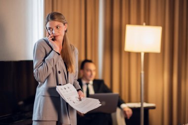 Klasik takım elbiseli güzel bir iş kadını cep telefonuyla konuşuyor, bazı verileri kontrol ediyor. Mavili meslektaşı arka planda oturuyor, bir koltukta oturuyor, dizüstü bilgisayarı üzerinde çalışıyor.