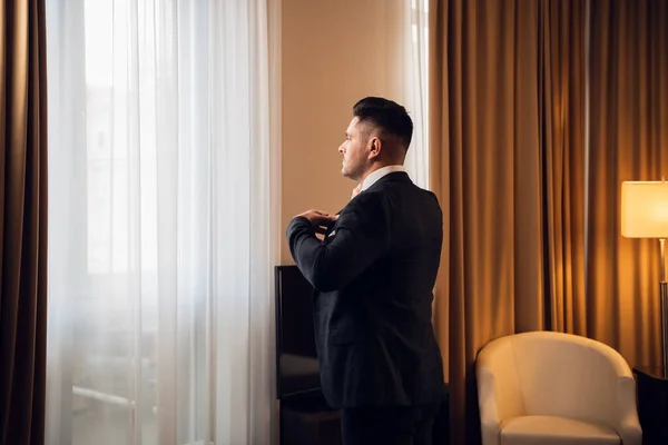 Un joven hombre de negocios guapo vestirse para una reunión, de pie frente a la ventana en un dormitorio — Foto de Stock