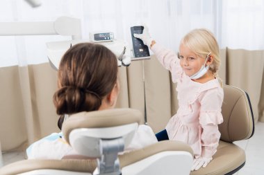 Küçük bir kız, dişçi kliniğinde oturmuş gülümserken ofisinde kadın bir pediatrik diş hekimi ile doktorculuk oynuyor.