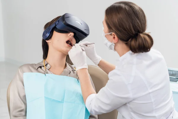 Dentystka wykonuje swoją pracę, podczas gdy pacjentka leży z otwartymi ustami, w okularach VR — Zdjęcie stockowe