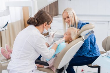 Annesiyle dişçi randevusunda olan bir çocuk. Kız ağzı açık, annesi arkada sandalyeye uzanıyor. Doktor işini yapıyor.