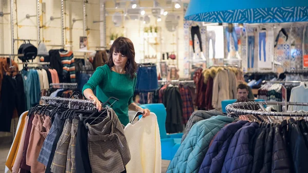 Женщина выбирает модную одежду в розничном магазине в торговом центре, в то время как ее мужчина ждет ее в гостиной, концепция продаж — стоковое фото