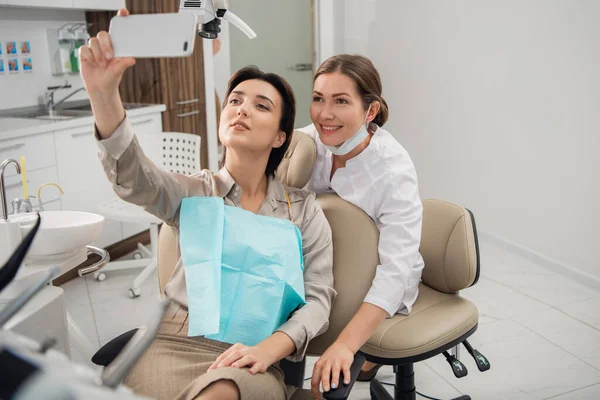 Νεαρή όμορφη ιατρός, οδοντίατρος, υγιεινολόγος ή ορθοδοντικός και ο ασθενής της βγάζει selfie με smartphone μετά από μια επιτυχημένη θεραπεία — Φωτογραφία Αρχείου