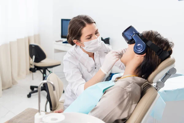Dentystka dokładnie badająca zęby swoich pacjentów, podczas gdy pacjent relaksuje się przy użyciu okularów VR — Zdjęcie stockowe