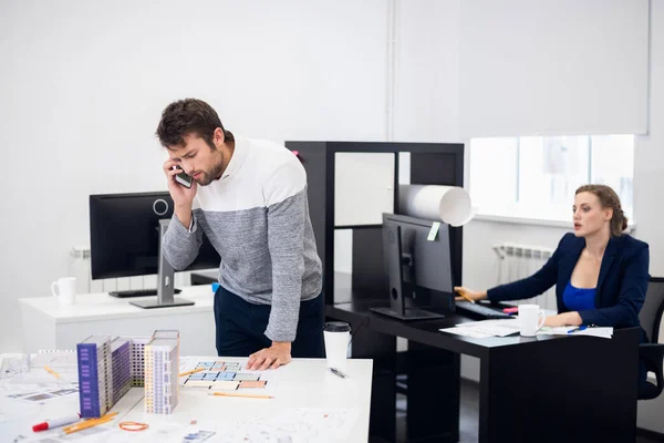 Ein Bauingenieur löst am Telefon einige Fragen, während seine Kollegin im Hintergrund an ihrem Computer arbeitet. — Stockfoto