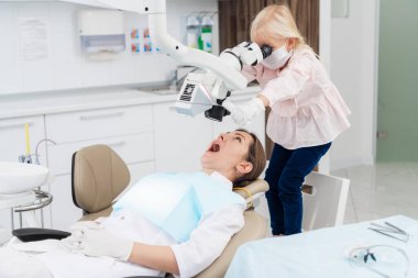 Özel ekipmanlarla sıhhiyecilerin ağzına bakan sevimli küçük kız. Doktor dişçide oturuyor ve ağzını sonuna kadar açık tutuyor. Çocuklar doktorculuk oynuyor. Dişçi ve