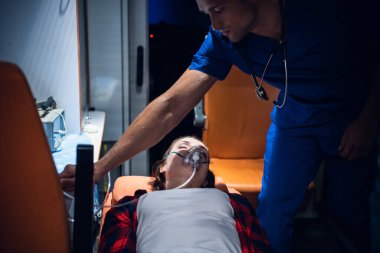 Genç bir kadın oksijen maskesi takmış sedyede yatıyor. Erkek bir sağlık görevlisi işini yapıyor.