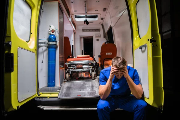 Un paramédico confuso con uniforme azul sentado en la parte trasera de un coche de ambulancia —  Fotos de Stock