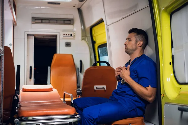 Уставший парамедик в синей форме держит стетоскоп, сидит в машине скорой помощи — стоковое фото