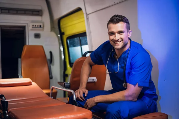 Um retrato de um homem sorridente com um uniforme médico azul sentado na ambulância — Fotografia de Stock