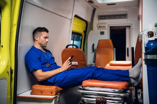 Мужчина парамедик в синей форме отдыхает и просматривает свой телефон в машине скорой помощи — стоковое фото