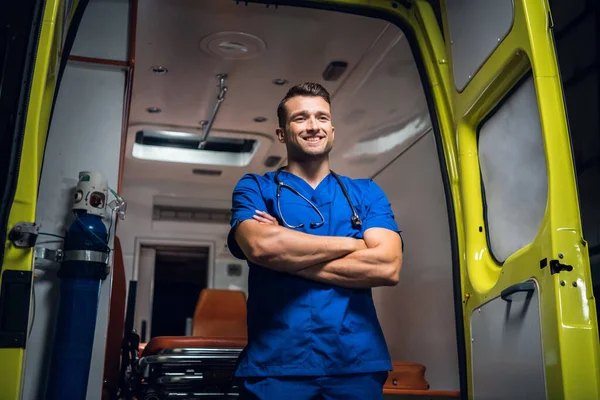 Joven médico de pie y sonriendo delante de un coche ambulancia —  Fotos de Stock