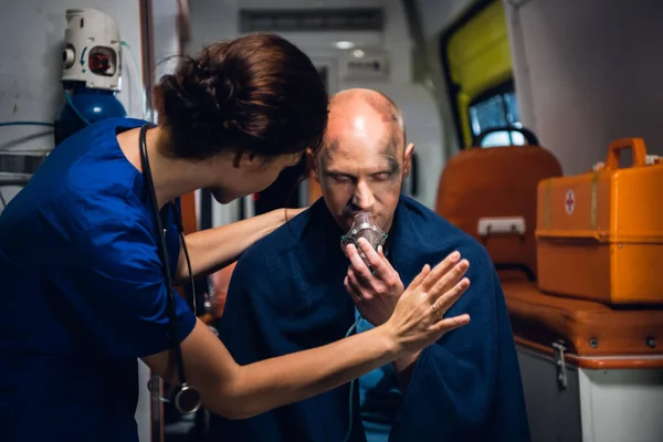 Una mujer joven con uniforme médico de pie con un hombre, que está sentado con una máscara de oxígeno en un coche de ambulancia —  Fotos de Stock