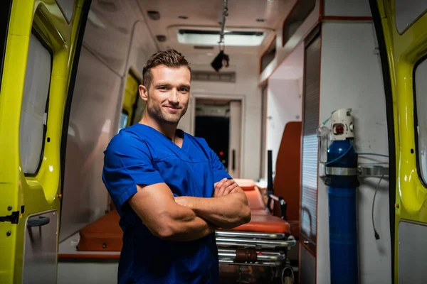 Junger Rettungssanitäter in Arztuniform blickt in die Kamera und lächelt, Krankenwagen im Hintergrund — Stockfoto