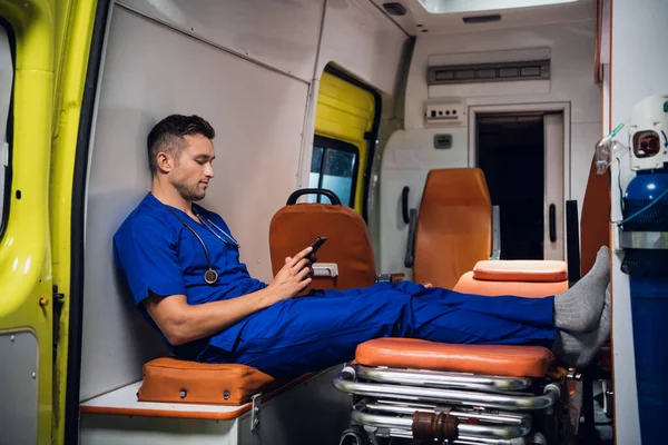 Молодой санитар сидит со своим телефоном в машине скорой помощи — стоковое фото