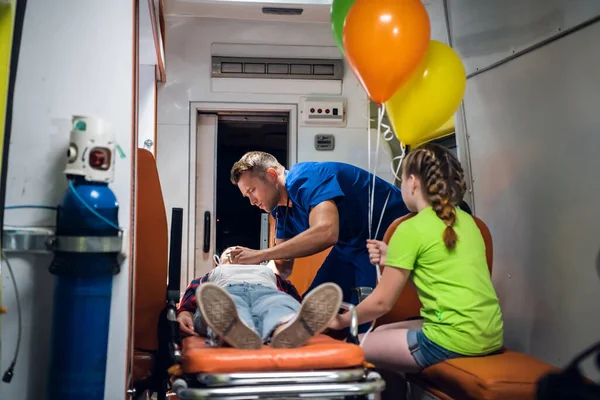 Молодой парамедик ухаживает за женщиной, лежащей на носилках в машине скорой помощи, пока ее маленькая дочь наблюдает за ними — стоковое фото