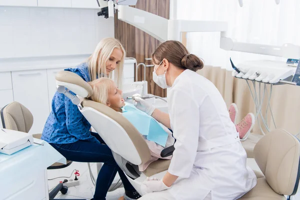 Uma criança com uma mãe numa consulta de dentista. A menina se deita na cadeira com a boca aberta, com a mãe para trás. O médico está a fazer o trabalho dela. — Fotografia de Stock