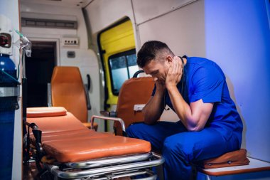 Ambulans arabasında oturan mavi üniformalı yorgun doktor.