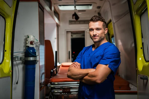 Tıbbi üniformalı yakışıklı sağlık görevlisi kameraya bakıyor ve gülümsüyor, arkada ambulans arabası var.