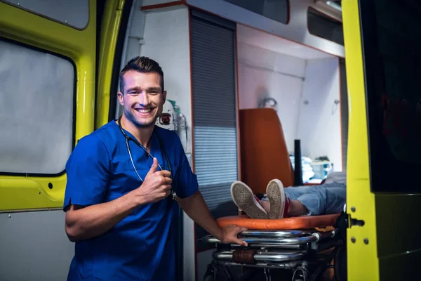 Un joven paramédico con uniforme azul se lleva a su paciente en una ambulancia, sonriendo y mostrando los pulgares hacia arriba, para animar a todos —  Fotos de Stock