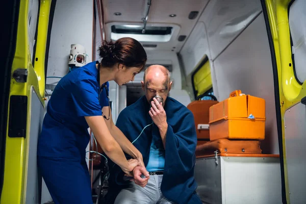 Раненый шокированный мужчина, сидящий с кислородной маской в машине скорой помощи, женщина в медицинской форме проверяет его пульс — стоковое фото