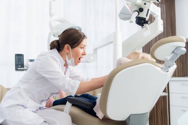 Küçük hastalarının dişlerini inceleyen genç bir dişçi ağızlarını daha geniş açmasını ve nasıl yapılacağını göstermesini istiyor.