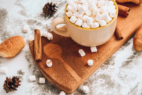 Чашка новогоднего вкусного горячего шоколада и какао с зефиром, посыпанного какао порошком, шишками и круассанами, корицей на сером столе, избирательный фокус — стоковое фото