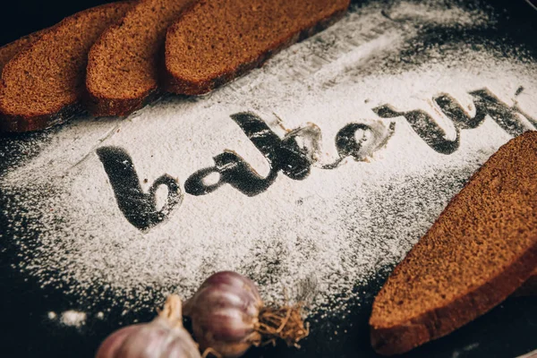 Inscripción panadería sobre harina de trigo blanco dispersa Pan de centeno rebanado con ajo sobre fondo oscuro — Foto de Stock