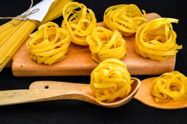 Çiğ spagetti ve makarna yuvarlak makarna topları. Siyah arka plandaki tahta kesme tahtasının üzerinde tahta kaşık var. İtalyan mutfağı..