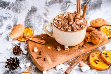 Tahta tahtaya kakao tozu serpiştirilmiş lezzetli bir fincan yeni yıl çikolatası ve kakao, kaşık şekli, portakal dilimleri, koniler ve kruvasanlar gri bir masada tarçın, seçici bir odak noktası, sıcak içecek kışı ya da