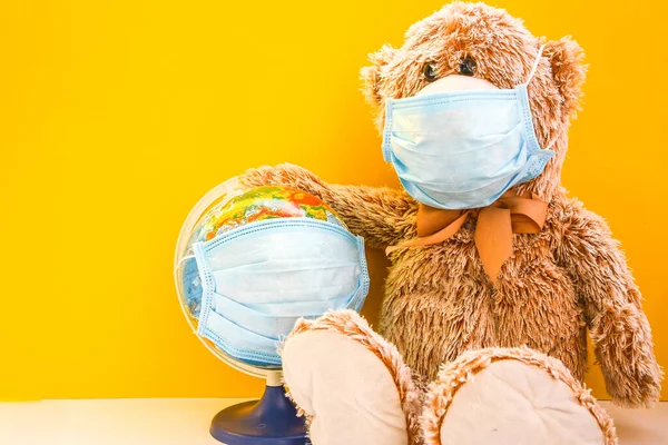 Juguete oso de peluche con máscara facial médica y la celebración del mundo de la tierra globo con máscara protectora sobre fondo amarillo, espacio de copia para su texto — Foto de Stock