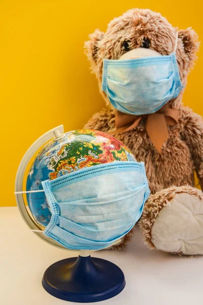 teddy bear toy wearing medical face mask and holding earth world globe with protective mask on yellow background, copy space for your text