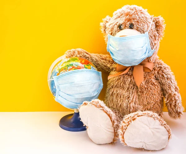 teddy bear toy wearing medical face mask and holding earth world globe with protective mask on yellow background, copy space for your text, quarantine against coronavirus, covid-19