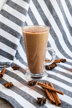 Sütlü, tarçınlı ve anason yıldızlı cam bardakta geleneksel kış içkisi gri bir bezin üzerinde krem şanti, kumaş arka planında tarçın çubukları. Kafe menüsü reklamı. Kahve.