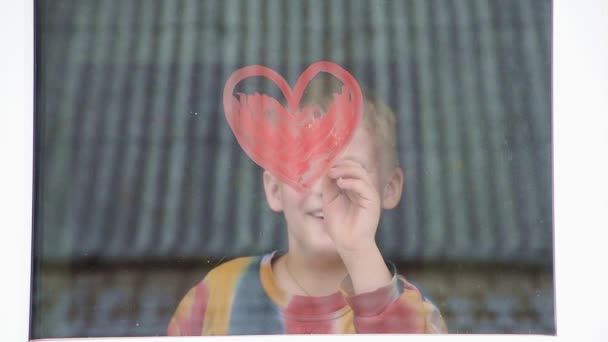 Quarantaine. De jongen tekent rood hart op het raam met verf. Wachten op de regenboog, thuisblijven — Stockvideo