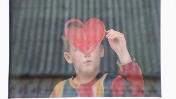 Cuarentena. El niño dibuja el corazón rojo en la ventana con pintura. Esperando el arco iris, Quédense en casa — Vídeo de stock