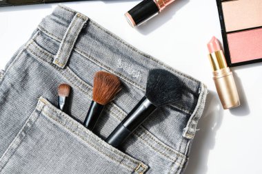 Farklı makyaj ürünleri kompozisyonu beyaz arka planda kot pantolon, düz yatış, üst görünüm, göz farı, ruj, fosforlu kalem, kot pantolon cebinde makyaj fırçası