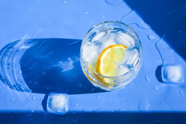 Mavi arka planda buzlu ve limonlu bir bardak ferahlatıcı su. Sıcaklık konsepti, taze. Doğal ışık. Düz konum, üst manzara, kopyalama alanı, cam bardakta soda.