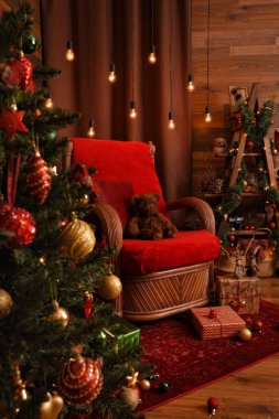 Kırmızı ve kahverengi gölgeli, koltuklu ve süslü Noel ağacıyla modern bir iç mekan. Sandalyede oturan oyuncak ayı, hediyeler, çelenkler ve diğer Noel süsleri.. 