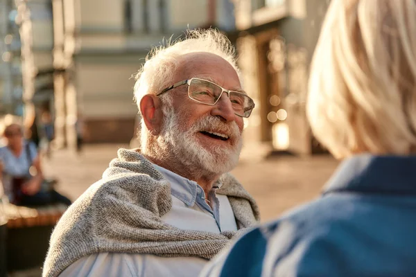 Счастливый старший бородатый мужчина в очках, смотрящий на жену и улыбающийся, сидя на скамейке вместе — стоковое фото