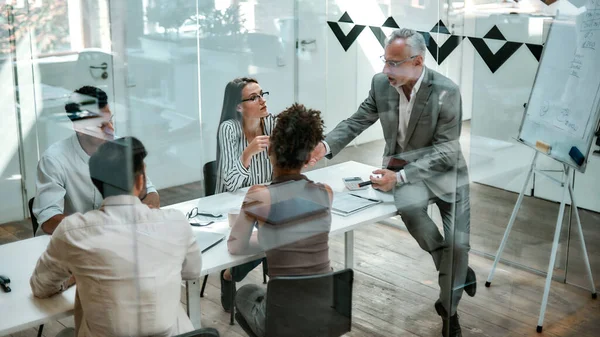 Zakenmensen. Zelfverzekerde volwassen man in formele kleding legt iets uit aan zijn collega 's terwijl hij op de kantoortafel zit achter de glazen muur in het moderne kantoor — Stockfoto