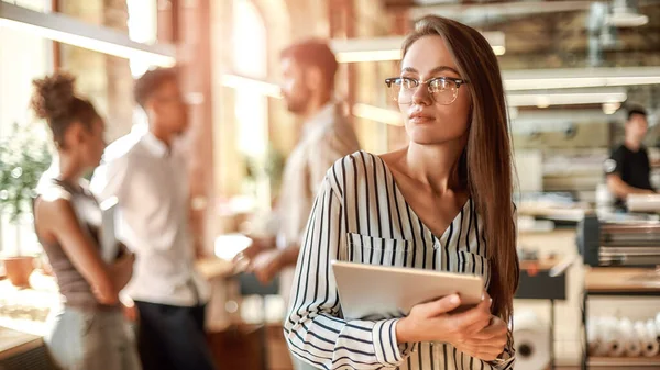 Smart och självsäker. ung kvinna i glasögon håller digital tablett och tittar på kameran när du står i det moderna kontoret — Stockfoto