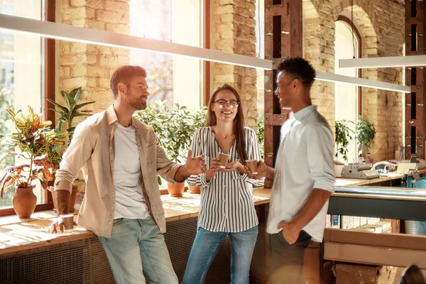 Отличный перерыв на кофе. Трое молодых и веселых коллег в повседневной одежде держат кофейные чашки и обсуждают что-то, стоя в современном офисе — стоковое фото