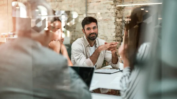 Jeune expert en affaires. Un homme souriant discute de quelque chose avec ses collègues assis à la table du bureau derrière le mur de verre — Photo