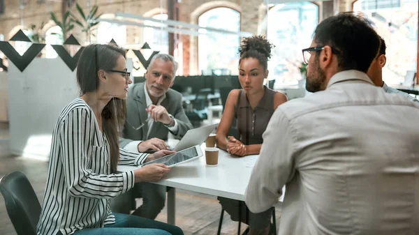 Usando tecnologías digitales. Joven mujer de negocios sosteniendo la tableta digital y discutiendo algo con sus colegas mientras están sentados en la mesa de la oficina juntos — Foto de Stock